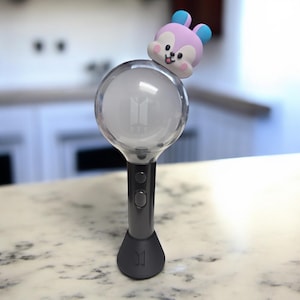 May include: A gray and black light stick with a clear, spherical head featuring the BTS logo. A small, cartoonish character with pink and blue accents sits atop the sphere. The light stick has two buttons and a flared base.