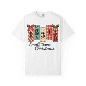 May include: White t-shirt featuring a festive design for Christmas. The design includes five rectangular gift boxes with numbers 1-5, each with a unique pattern and bow. The text "Small town Christmas" is printed below the boxes.