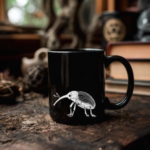 以下が含まれることがあります： 白いゾウムシのイラストが描かれた黒いセラミックマグ。 昆虫は長い鼻と縞模様の体を持っています。 マグカップには湾曲したハンドルが付いています。 背景はぼやけています。