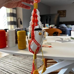 May include: A handmade macrame plant hanger in white, orange, and yellow, holding a ceramic seagull figurine wearing a sailor hat. The seagull has a yellow beak and feet. The macrame has a braided top and a tassel at the bottom.