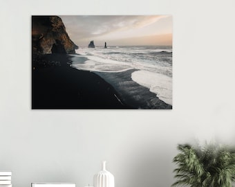 May include: A landscape photograph showing a black sand beach with waves breaking on the shore. The sky is a blend of orange and grey, and distant rock formations are visible.