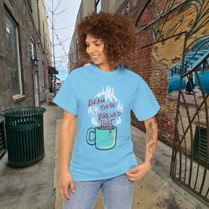 May include: A light blue short-sleeve t-shirt featuring a graphic of a teal coffee mug with steam rising above it. The text "BEAN THERE BREWED THAT" is printed in dark red letters within the steam design on the front of the garment.