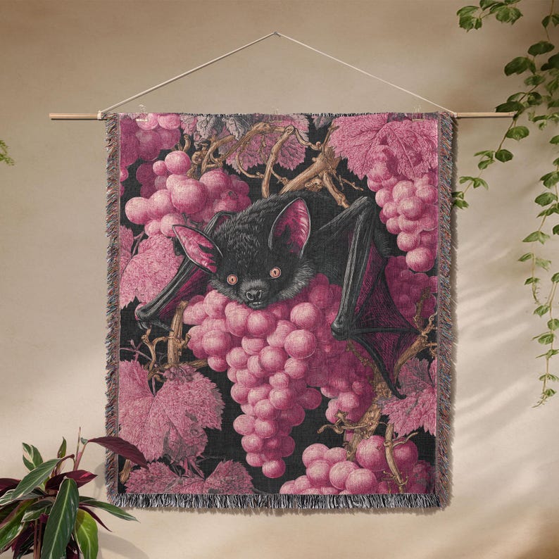 May include: A wall hanging featuring a black bat with red accents, surrounded by pink grapes and leaves against a black background. The tapestry is fringed at the bottom and suspended from a wooden rod.