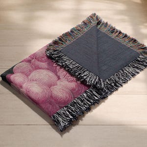 May include: A folded throw blanket with a pink floral pattern and a gray reverse side. The blanket has a fringe border with a mix of colors. The floral design features a cluster of pink, round shapes.