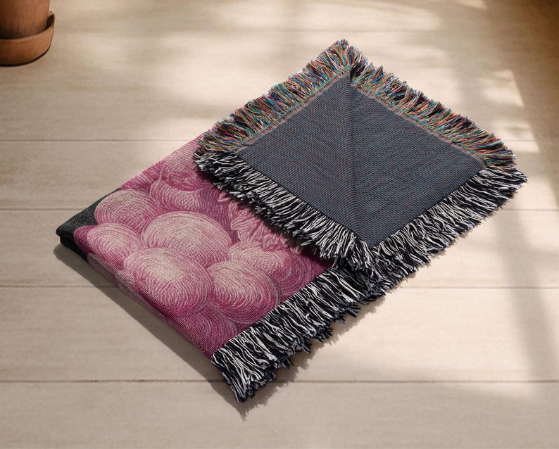May include: A folded throw blanket with a pink floral pattern and a gray reverse side. The blanket has a fringe border with a mix of colors. The floral design features a cluster of pink, round shapes.