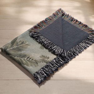 Pu&ograve; includere: Una coperta piegata con un motivo a foglie botaniche nei toni del verde e del grigio. La coperta ha un retro blu scuro ed &egrave; rifinita con una frangia multicolore. &Egrave; appoggiata su una superficie di legno chiaro.