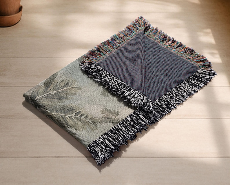 Pu&ograve; includere: Una coperta piegata con un motivo a foglie botaniche nei toni del verde e del grigio. La coperta ha un retro blu scuro ed &egrave; rifinita con una frangia multicolore. &Egrave; appoggiata su una superficie di legno chiaro.