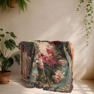 K&ouml;nnte beinhalten: Eine floral gemusterte Tagesdecke, die &uuml;ber einem Korbstuhl drapiert ist. Die Decke zeigt gro&szlig;e rosa und rote Rosen auf einem teal-farbenen Hintergrund mit einem Fransenrand. Der Stuhl steht in einem Raum mit Pflanzen.