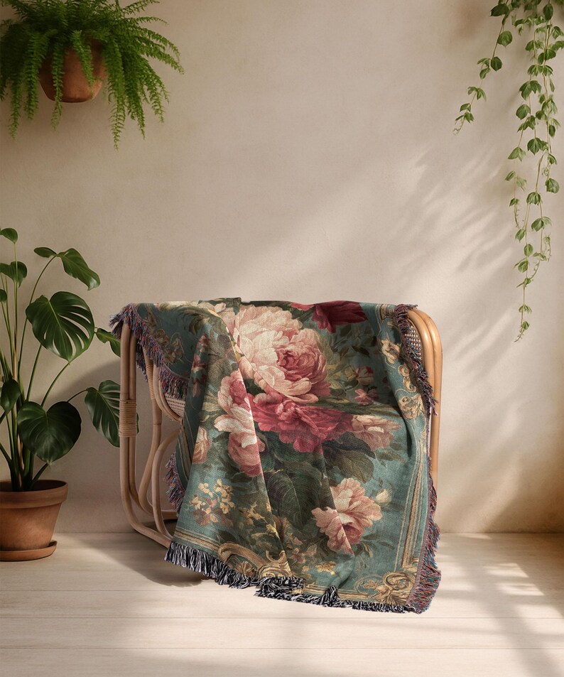 K&ouml;nnte beinhalten: Eine floral gemusterte Tagesdecke, die &uuml;ber einem Korbstuhl drapiert ist. Die Decke zeigt gro&szlig;e rosa und rote Rosen auf einem teal-farbenen Hintergrund mit einem Fransenrand. Der Stuhl steht in einem Raum mit Pflanzen.