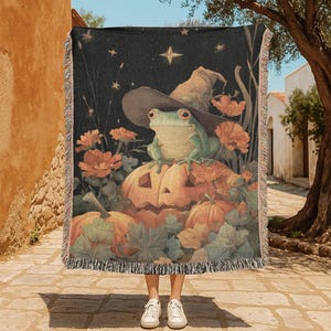 Op de afbeelding: Een geweven sprei met een Halloween-thema. Het ontwerp toont een groene kikker met een heksenhoed op een uitgesneden pompoen. De achtergrond is een donkere nachthemel met sterren en bloemen. De sprei heeft een gefranjerde rand.