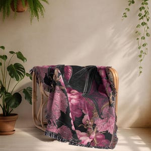 May include: A woven throw blanket draped over a wicker chair. The blanket features a dark background with a large floral pattern in shades of pink, purple, and black. The edges are finished with a fringe.