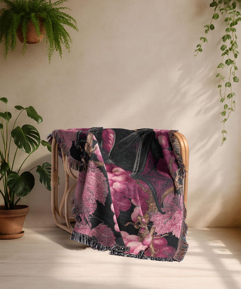 May include: A woven throw blanket draped over a wicker chair. The blanket features a dark background with a large floral pattern in shades of pink, purple, and black. The edges are finished with a fringe.
