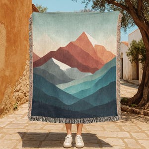 May include: A woven throw blanket featuring a mountain landscape design in shades of blue, rust, and white. The blanket has a fringed edge and is held up in front of a person. The design depicts a stylized mountain range.