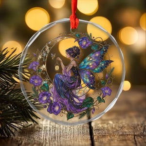 May include: A circular, faceted glass ornament featuring a printed illustration of a fairy with iridescent wings, sitting within a crescent moon shape surrounded by purple morning glory flowers and foliage, suspended by a red ribbon.