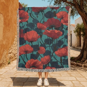 Op de afbeelding: Een rechthoekige geweven plaid met een herhalend patroon van grote rode klaprozen met donkergroene stengels en bladeren tegen een groenblauwe achtergrond. De deken heeft een decoratieve franjerand aan de boven- en onderkant.