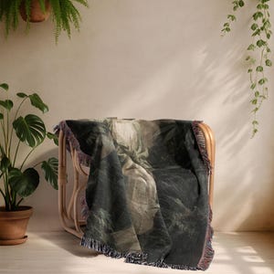 May include: A dark green and beige throw blanket with a botanical print is draped over a light brown wicker chair. The blanket has a fringed edge with hints of purple and red. The scene is set in a room with plants.