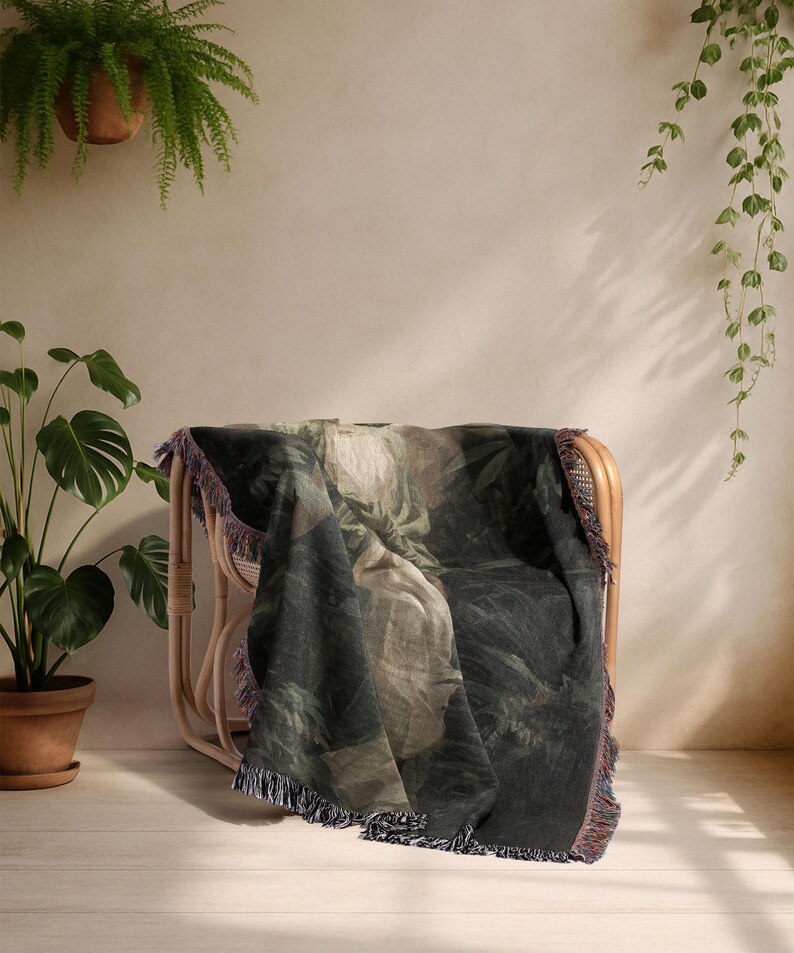 May include: A dark green and beige throw blanket with a botanical print is draped over a light brown wicker chair. The blanket has a fringed edge with hints of purple and red. The scene is set in a room with plants.