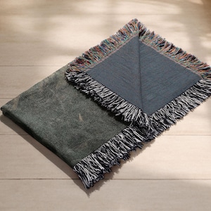 May include: A folded, dark green throw blanket with a textured pattern and fringed edges. The blanket is partially unfolded, revealing a section with a blue-grey background and colorful fringe. The blanket is on a light wood surface.