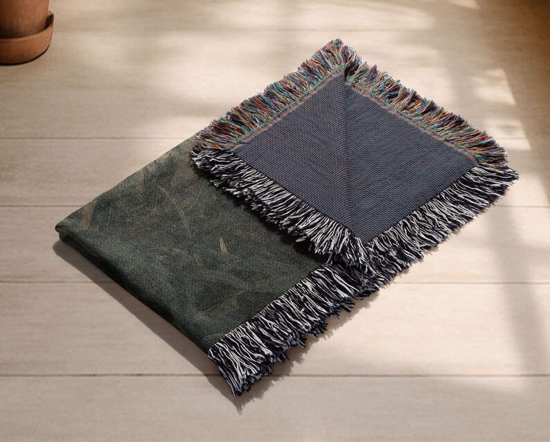 May include: A folded, dark green throw blanket with a textured pattern and fringed edges. The blanket is partially unfolded, revealing a section with a blue-grey background and colorful fringe. The blanket is on a light wood surface.