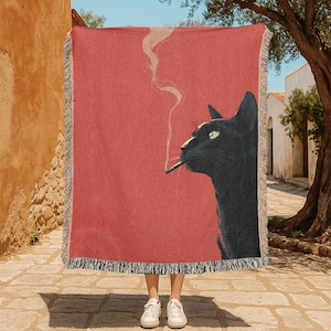 May include: A red throw blanket featuring a black cat smoking a cigarette. The cat is depicted in profile with green eyes and smoke rising from the cigarette. The blanket has a fringed edge.