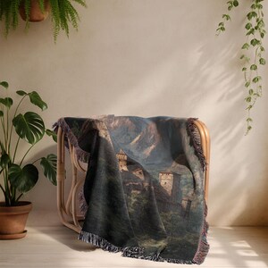 May include: A woven throw blanket with a castle landscape design, draped over a rattan chair. The blanket's colours include green, blue, and brown. The edges are finished with a fringe. The chair is made of rattan.