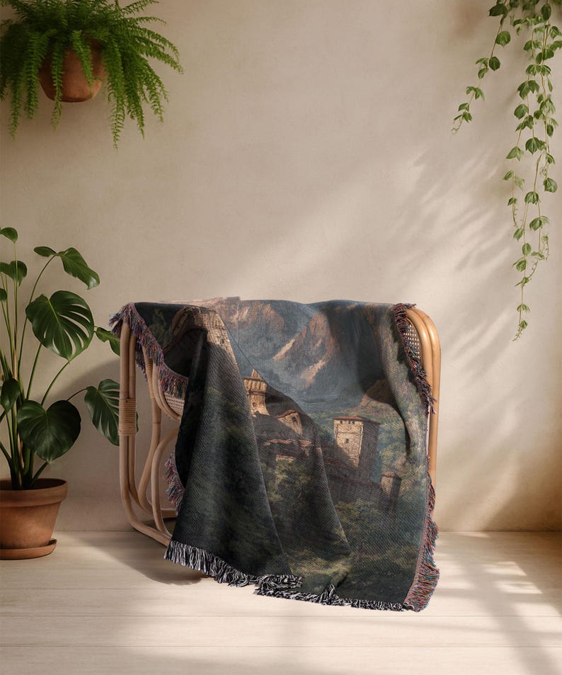 May include: A woven throw blanket with a castle landscape design, draped over a rattan chair. The blanket's colours include green, blue, and brown. The edges are finished with a fringe. The chair is made of rattan.