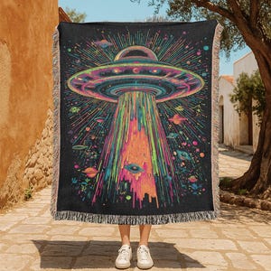 May include: Black throw blanket with a vibrant UFO design, emitting a beam of light. The illustration includes planets and stars in pink, blue, green, and orange. The blanket has a fringed edge.