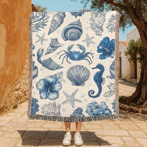 May include: A cream-colored throw blanket featuring a blue nautical design. The pattern includes seashells, starfish, coral, a seahorse, and hibiscus flowers. The blanket has a fringed edge and is held up in front of a person.