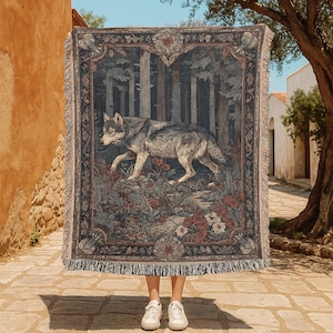 Peut inclure: Un plaid tissé présentant une illustration détaillée d'un loup gris marchant dans une forêt dense avec des fleurs rouges et blanches au premier plan. La couverture possède une bordure florale décorative et des franges en haut et en bas.