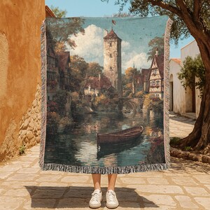May include: Woven throw blanket featuring a detailed landscape painting of a European town with a river, bridge, and tower. The artwork has a vintage aesthetic with muted colors and a fringed edge. The blanket is held up by a person wearing white sneakers.