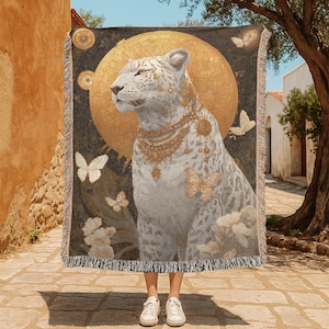May include: A woven throw blanket featuring a white leopard with gold jewelry and a golden halo. The design includes butterflies and floral elements on a dark background with a fringed edge. The blanket is held up by a person wearing white sneakers.