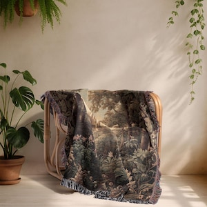 Op de afbeelding: Een geweven sprei met een gedetailleerde bossc&egrave;ne, gedrapeerd over een rotan stoel. De deken heeft een gefranjerde rand en is in tinten groen, bruin en beige. De stoel staat tegen een neutrale muur, met hangende planten.