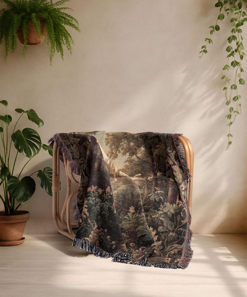 Op de afbeelding: Een geweven sprei met een gedetailleerde bossc&egrave;ne, gedrapeerd over een rotan stoel. De deken heeft een gefranjerde rand en is in tinten groen, bruin en beige. De stoel staat tegen een neutrale muur, met hangende planten.