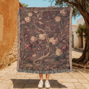 May include: A woven throw blanket with a floral tapestry design in shades of burgundy, cream, and green. The blanket features a detailed pattern of flowers, branches, and foliage, with a fringed edge. The blanket is held up in front of a person.