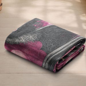May include: Folded blanket with a floral pattern in shades of gray, white, and magenta. The blanket is folded neatly, showcasing the design. The fabric appears soft and textured, suitable for home decor.