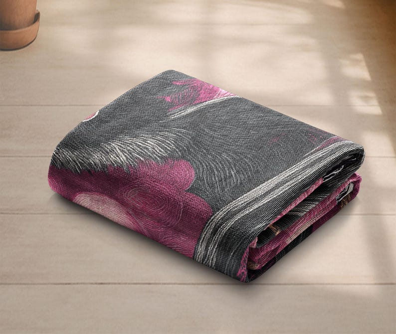 May include: Folded blanket with a floral pattern in shades of gray, white, and magenta. The blanket is folded neatly, showcasing the design. The fabric appears soft and textured, suitable for home decor.