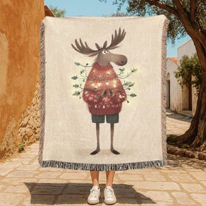 May include: A woven throw blanket with a cartoon moose wearing a red Christmas sweater and shorts. The moose is decorated with fairy lights. The blanket has a fringed edge and a neutral background. The blanket is suitable for a sofa or bed.