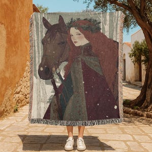 May include: A woven throw blanket featuring a detailed illustration of a woman with long red hair and a horse. The woman wears a burgundy cloak and a green wreath. The background shows a snowy forest scene. The blanket has a fringed edge.