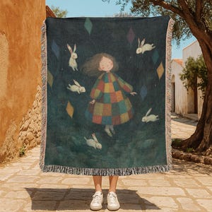 Può includere: Una coperta blu scuro con un'illustrazione stravagante di una ragazza in un abito a scacchi colorato circondata da conigli bianchi e forme di diamanti colorati. La coperta ha un bordo a frange.