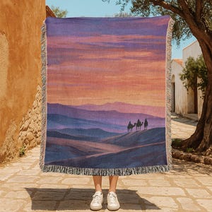 Pode incluir: Uma manta tecida com uma paisagem desértica com camelos e um pôr do sol em tons de roxo, laranja e azul. A manta tem uma borda com franjas. Uma pessoa com tênis brancos segura a manta.