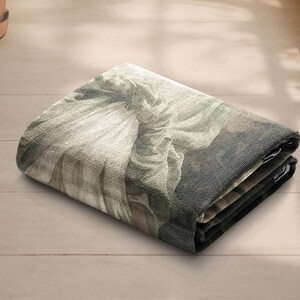 May include: Folded rectangular tapestry featuring a classical artwork print in shades of gray, beige, and brown. The tapestry is folded neatly, showcasing the detailed design and texture of the fabric. The artwork depicts a figure with flowing garments.
