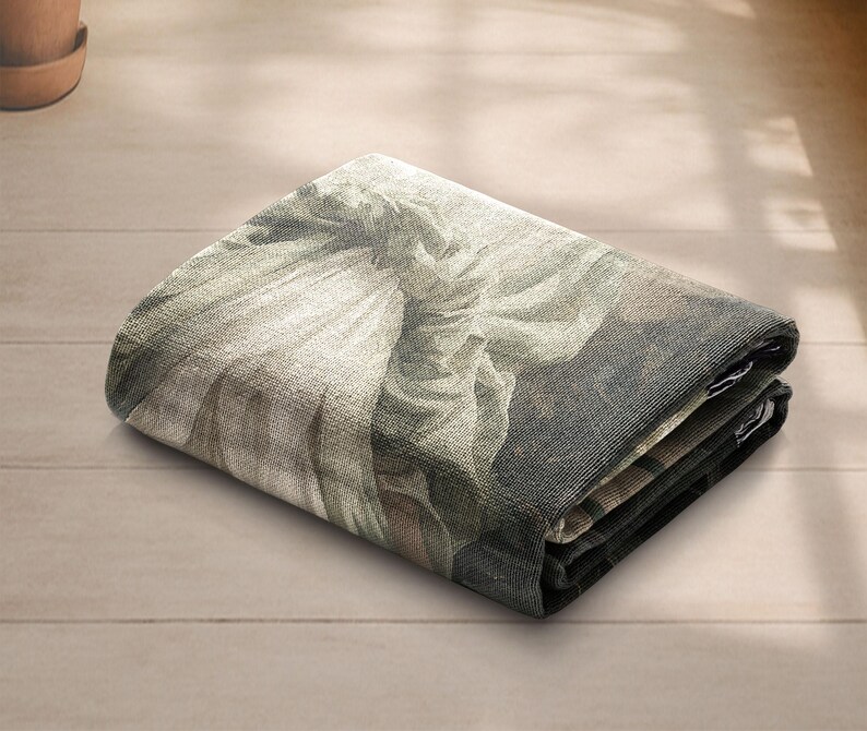 May include: Folded rectangular tapestry featuring a classical artwork print in shades of gray, beige, and brown. The tapestry is folded neatly, showcasing the detailed design and texture of the fabric. The artwork depicts a figure with flowing garments.