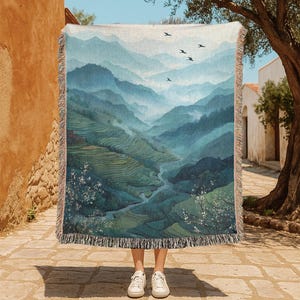 May include: A woven throw blanket featuring a scenic landscape of layered blue and green mountains, a river, and flying birds. The blanket has a fringed edge and is held up by a person wearing white sneakers.