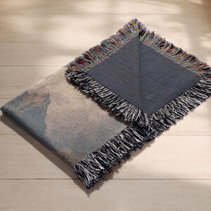 May include: A folded woven throw blanket featuring a landscape design in blue, beige, and brown hues. The blanket has a dark blue border and a multi-coloured fringe. It is displayed on a light wooden surface.