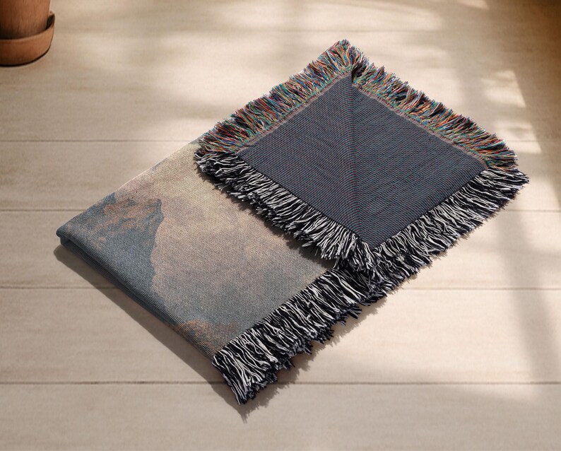 May include: A folded woven throw blanket featuring a landscape design in blue, beige, and brown hues. The blanket has a dark blue border and a multi-coloured fringe. It is displayed on a light wooden surface.