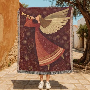 May include: A burgundy throw blanket featuring an angel playing a trumpet. The angel has gold wings and is wearing a red dress. The blanket has a fringe border and a pattern of gold stars and swirls.