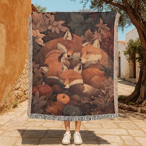 May include: A woven throw blanket depicting a group of foxes sleeping amongst autumnal foliage and pumpkins. The blanket, with a fringed border, uses a palette of brown, orange, and green, creating a warm and inviting aesthetic. The blanket is perfect for a sofa or bed.