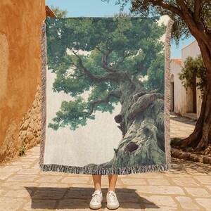Puede incluir: Una manta tejida rectangular que presenta una ilustración detallada de un árbol grande y nudoso con corteza texturizada y follaje verde exuberante. La manta tiene un borde con flecos y muestra una escena naturalista sobre un fondo claro.