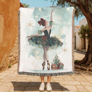May include: A woven throw blanket featuring a watercolor illustration of a ballerina in a dark green tutu, with a Christmas tree and presents. The blanket has a fringed edge and a light blue and white background.