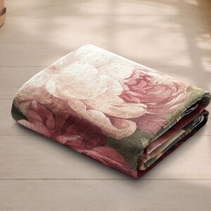 K&ouml;nnte beinhalten: Gefaltete Blumendecke mit einem gro&szlig;en Rosenmuster in Rosa, Creme und Gr&uuml;n. Die Decke ist ordentlich gefaltet und zeigt das detaillierte Design und die Textur des Stoffes. Die Decke liegt auf einer hellen Holzoberfl&auml;che.